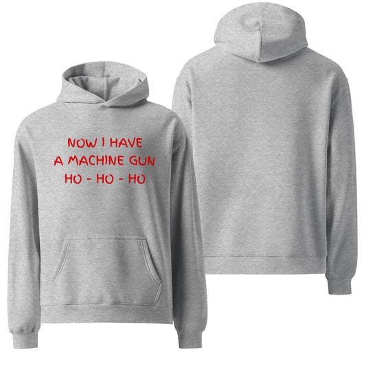 An oversized hoodie in athletic heather gray featuring the text "Now I have a machine gun, ho ho ho" in bold, red letters. The design references the iconic line from the movie Die Hard, blending holiday humor with a cult-classic vibe.