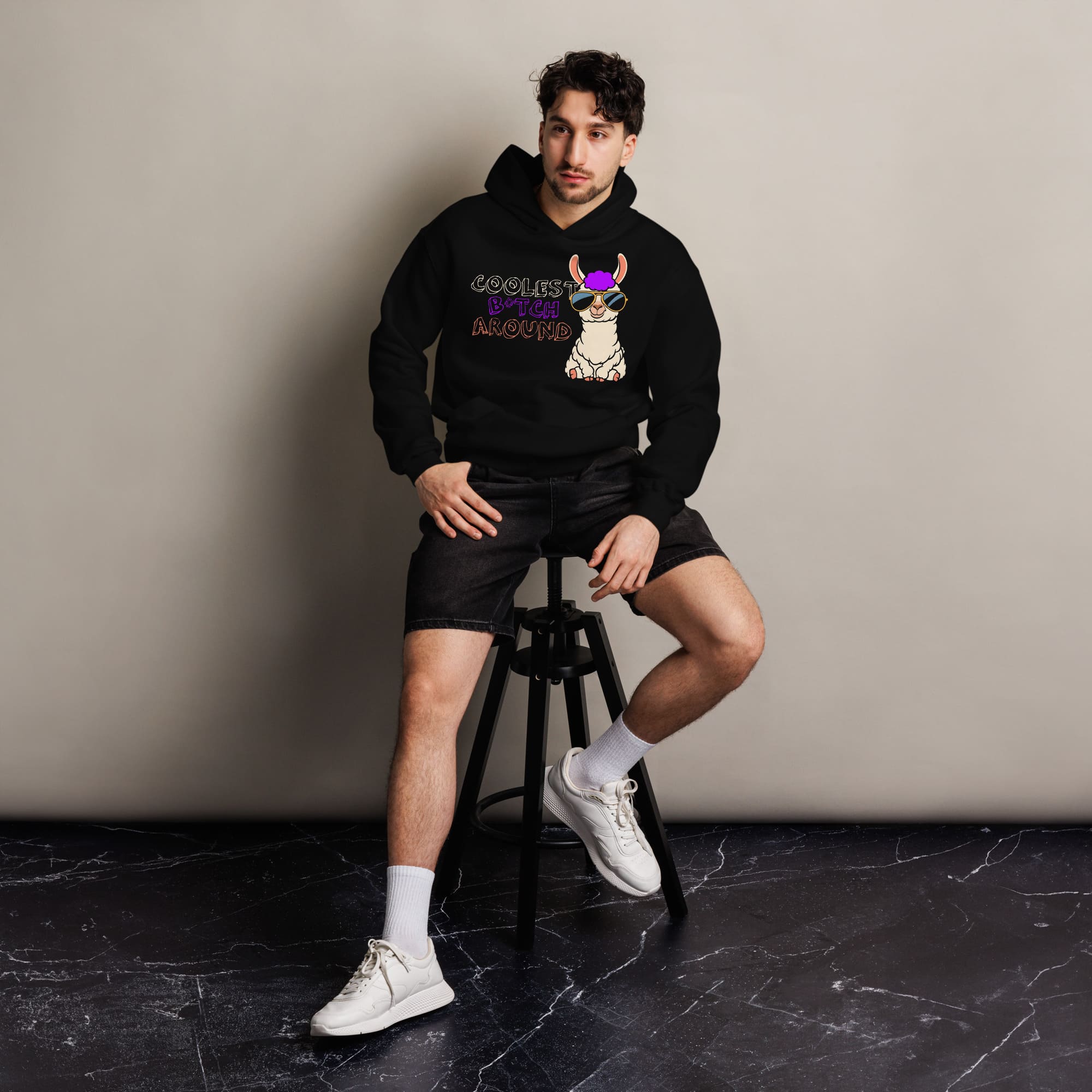 Person seated on a stool wearing a black hoodie with a colorful dog design, black shorts, white socks, and white sneakers, against a neutral background.